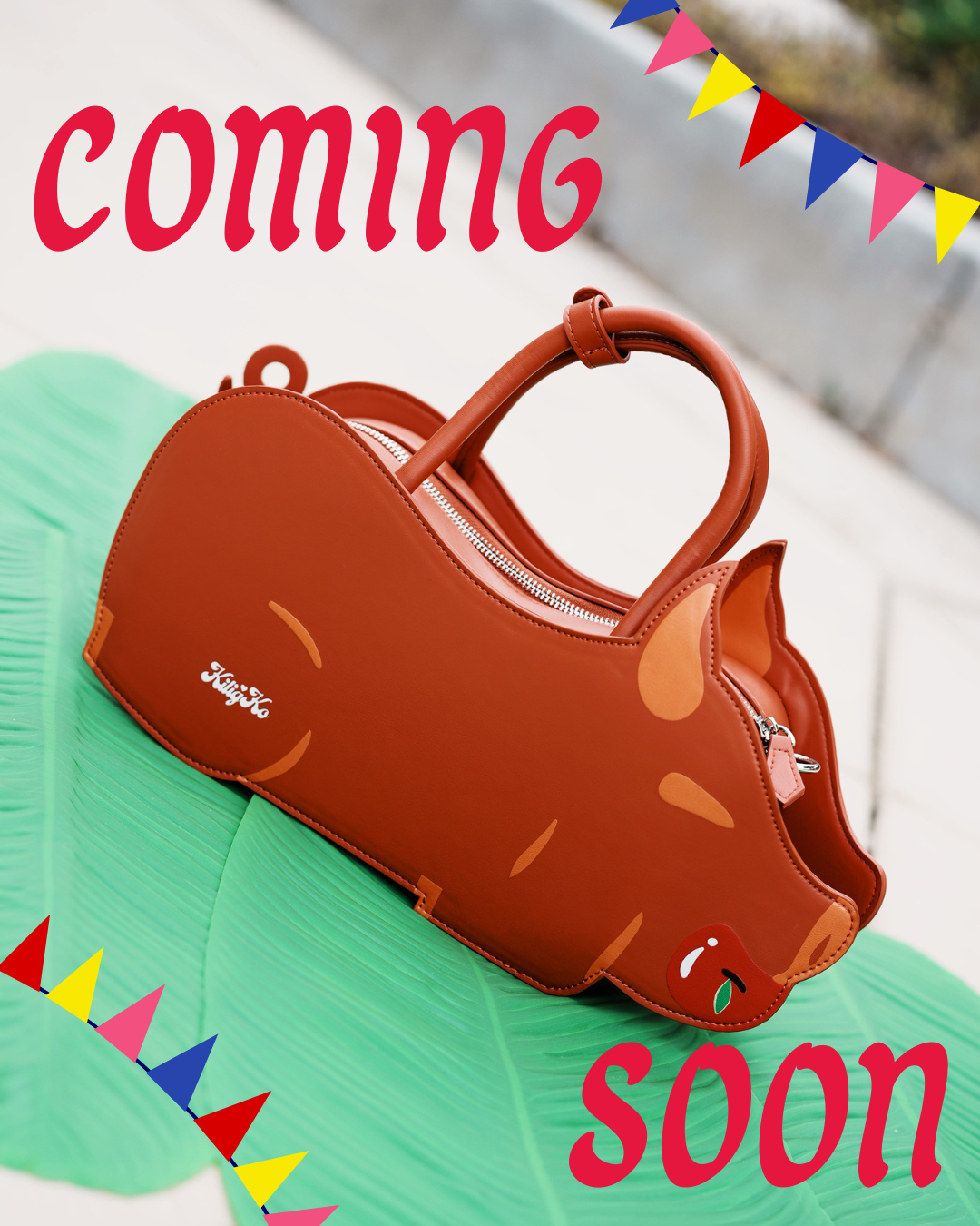 A brown lechon-shaped handbag with a top handle, shown on a green leaf-like mat with a blurred outdoor background. Red text at the top reads 'coming soon!'.