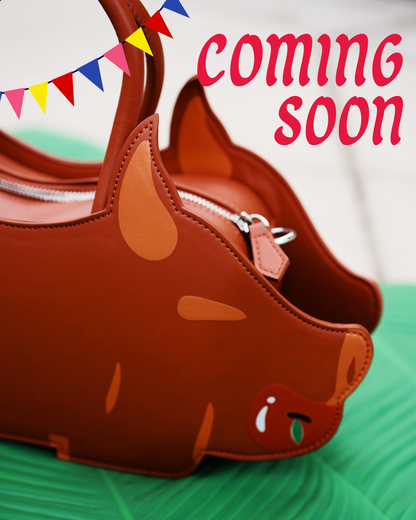 A close up shot of brown lechon-shaped handbag with a top handle, shown on a green leaf-like mat with a blurred outdoor background. Red text at the top reads 'coming soon!'.